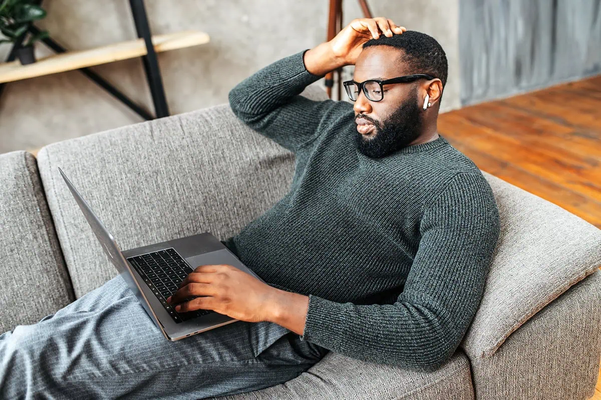 Maximum Pay, Minimum Mileage: 12 Lucrative Jobs You Can Do from Anywhere 2 a black man in his 30s rests on a couch while using his laptop.