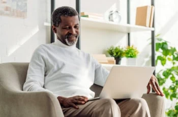 a black man wearing a white sweater smiles while looking at his laptop.