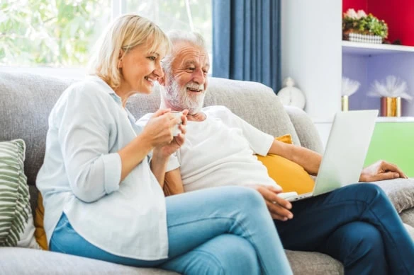 an older man and woman smile while drinking coffee and looking at their laptop.