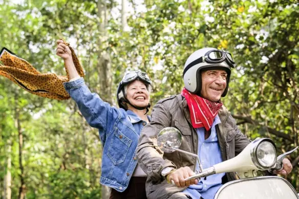 a senior couple smiles while riding on a scooter in the woods.