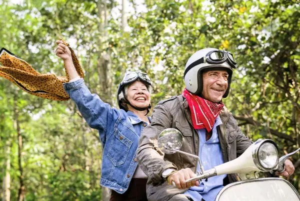 a senior couple smiles while riding on a scooter in the woods.