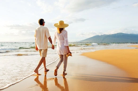 two older people walk on a sunny beach.