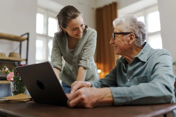 a senior man smiles at his adult daughter while working on his laptop.