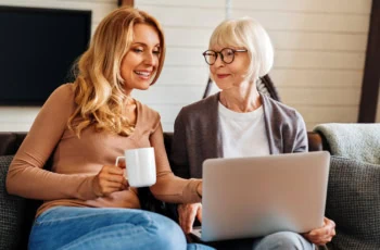 A happy senior mom with her adult daughter looking at a laptop while sitting on the couch.