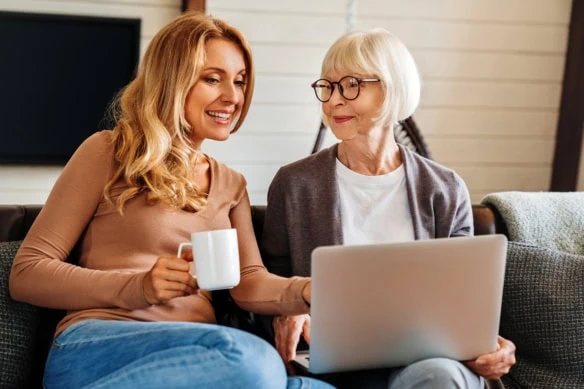 A happy senior mom with her adult daughter looking at a laptop while sitting on the couch.