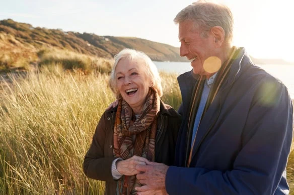 a loving active senior couple walk arm in arm through a field near a rocky shore.