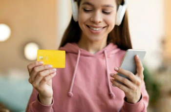 A girl in headphones looks at a smartphone and a debit card. 1200