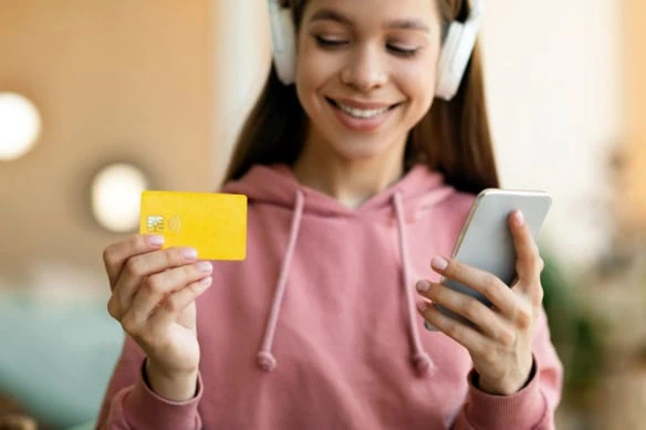 A girl in headphones looks at a smartphone and a debit card. 1200