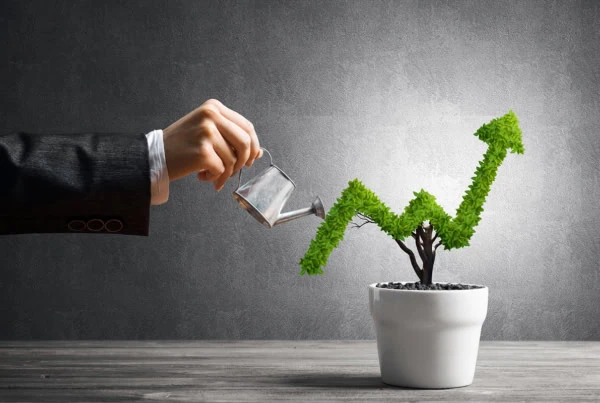 Hand of woman watering small plant in pot shaped like a growth arrow in a chart.