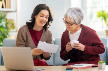 a middle-aged woman and her adult daughter look at financial statements while talking on the couch.