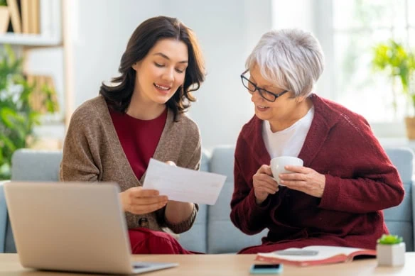 a middle-aged woman and her adult daughter look at financial statements while talking on the couch.