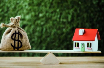 a bag of cash and a house sit on opposite sides of a seesaw.