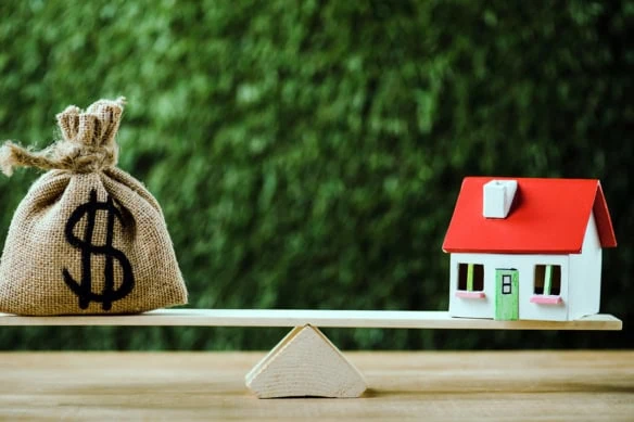 a bag of cash and a house sit on opposite sides of a seesaw.