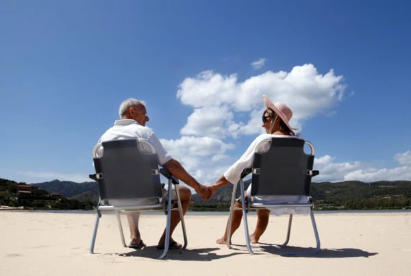 a senior couple holding hands while sitting on chairs at the beach.