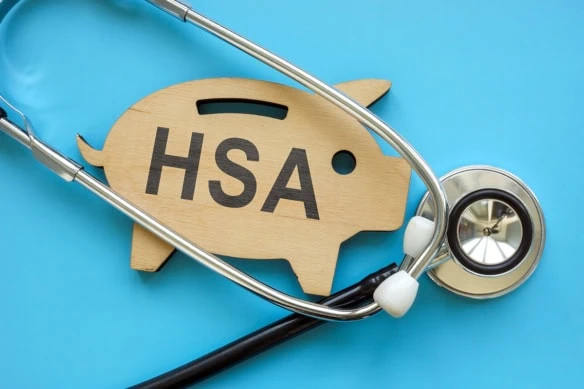 a wooden cutout of a piggy bank that says H S A sits against a blue background with a stethoscope.