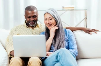 a happy senior couple looks at a computer while sitting at their couch.