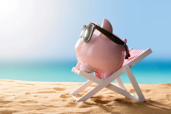 a piggy bank wears sunglasses and sits on a beach chair resting on the sand.