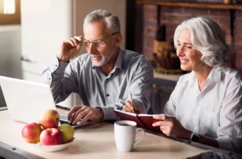 Senior couple sitting at the kitchen table looking at something on a laptop and smiling.