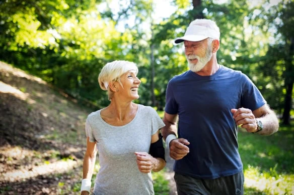 an older couple smiling at each other while jogging in a park.