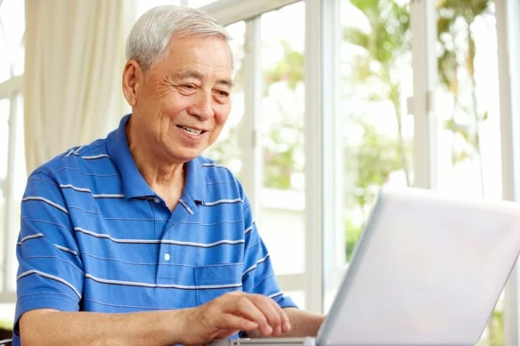 an older Chinese man smiles while looking at his laptop.