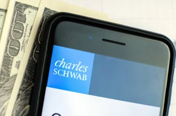 hundred dollar bills lying next to a smartphone displaying the charles schwab website.