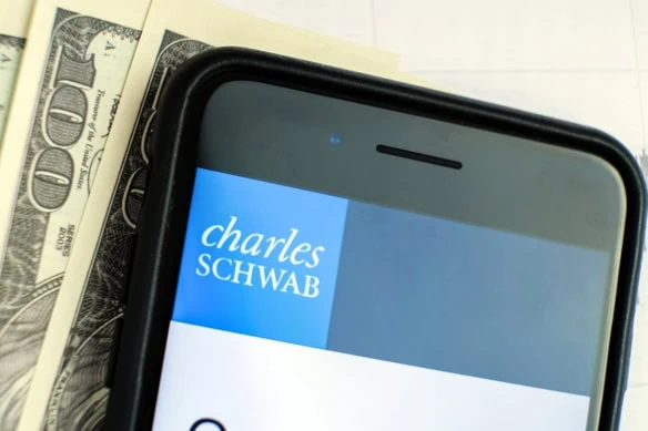 hundred dollar bills lying next to a smartphone displaying the charles schwab website.