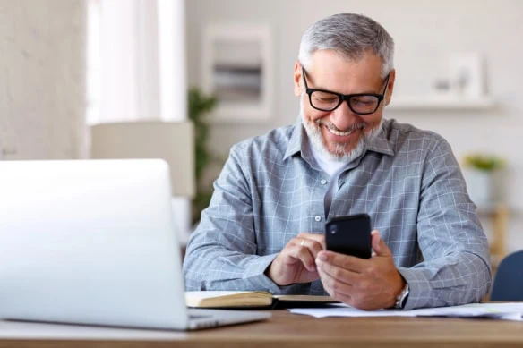 a man smiles while looking down at his smartphone at his desk at home.