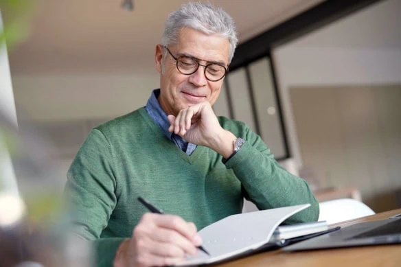 an older man wearing a green shirt looks at his finances on his computer and a notebook.