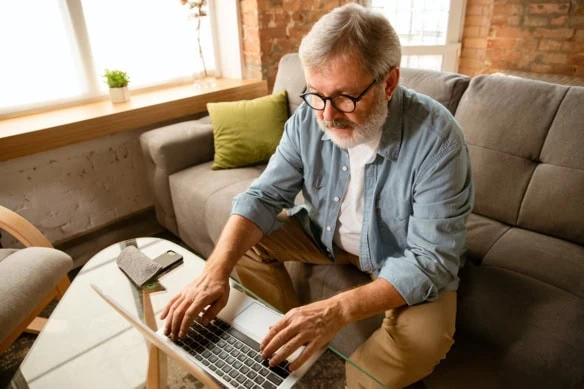 An older man is looking at his laptop while sitting on a couch.