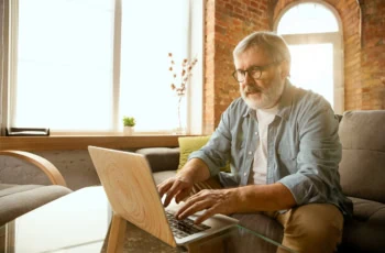 an older man works with his laptop at home in a sun-drenched room.