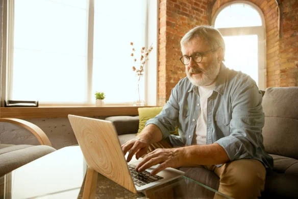 an older man works with his laptop at home in a sun-drenched room.