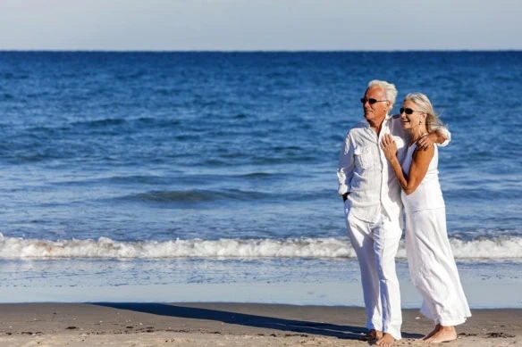 a well-dressed senior couple stands on the beach looking off into the distance.