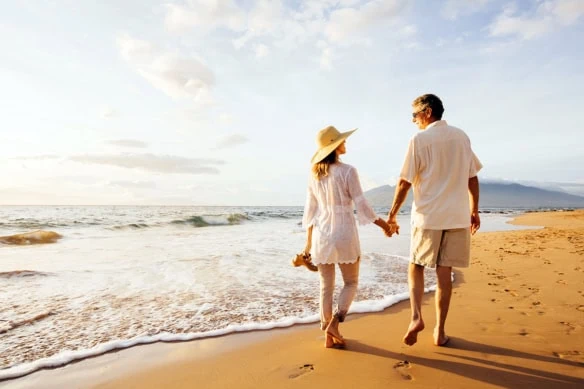 a retired couple walks barefoot on a beach on a sunny day.