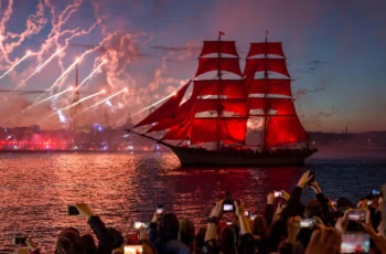 a ship with red sails moves along a river during a fireworks show.