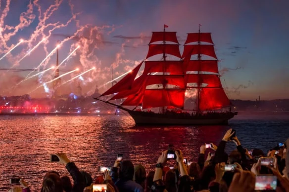 a ship with red sails moves along a river during a fireworks show.