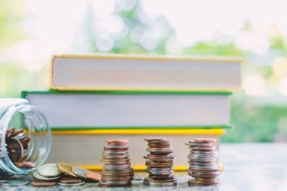 Pile of money coins outside the glass jar in front of books on blurred natural green background with books