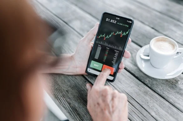 over-the-shoulder view of a woman using her smartphone to buy and sell stocks.