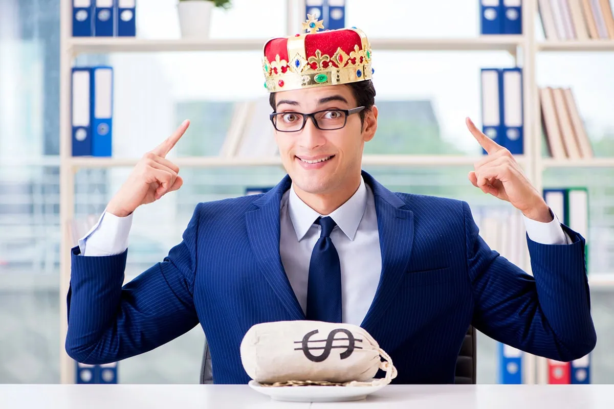 Get Income + Diversification With These 10 Top Dividend ETFs 5 a businessman sits at a desk with a bag of money on it and he points at the crown on his head.