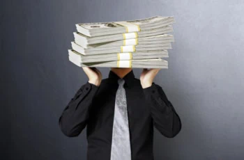 a man tries to lift stacks of comically large hundred dollar bills over his head.