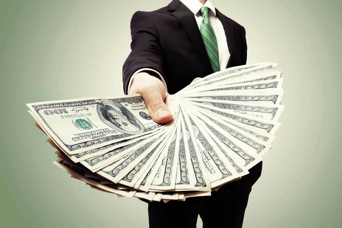a businessman handing out a fanned out stack of hundred dollar bills.