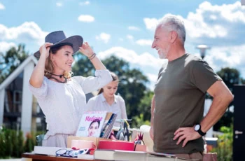 A smiling woman puts on a hat at a garage sale while an older man looks on.