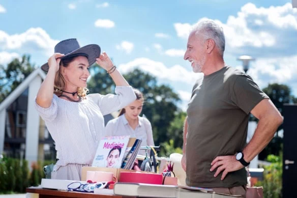 A smiling woman puts on a hat at a garage sale while an older man looks on.
