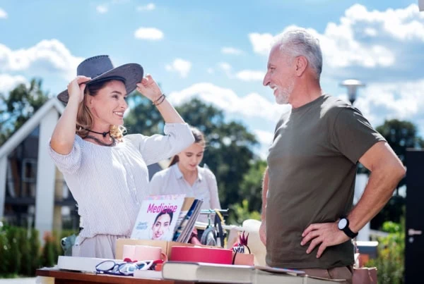 A smiling woman puts on a hat at a garage sale while an older man looks on.
