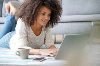 young generation z woman smiles while lying down and looking at her laptop.
