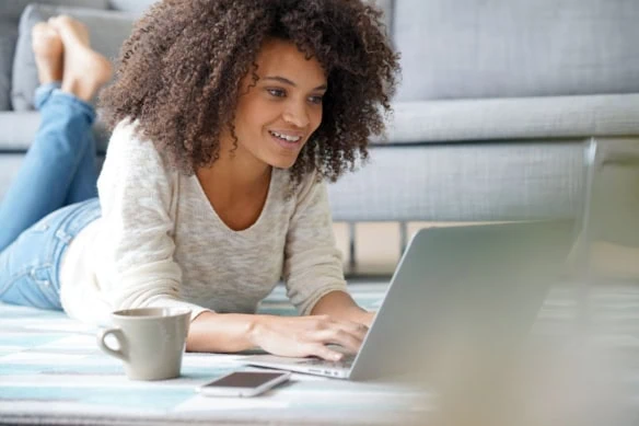 young generation z woman smiles while lying down and looking at her laptop.