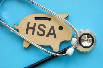 a wooden cutout of a piggy bank that says H S A sits against a blue background with a stethoscope.