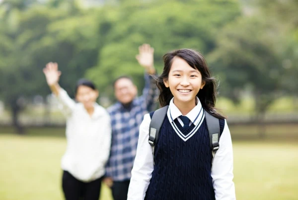 girl wearing backpack and parents waving in background