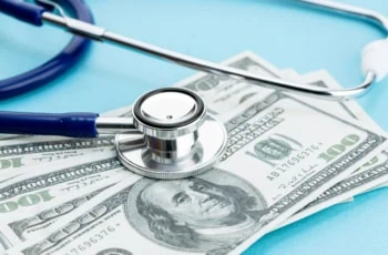 a stethoscope sits on hundred dollar bills sitting on a blue table.