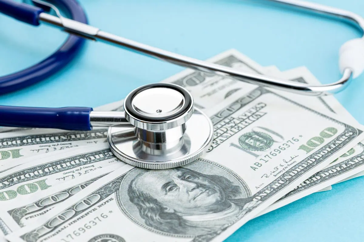 7 Fidelity Funds for a Healthier HSA in 2026 7 a stethoscope sits on hundred dollar bills sitting on a blue table.
