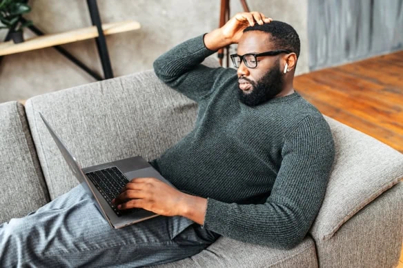 a black man in his 30s rests on a couch while using his laptop.